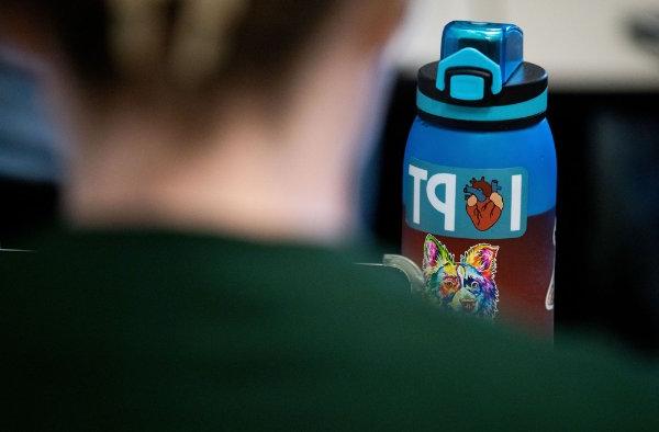 A water bottle has the word the letter "I," an image of an anatomical heart, then the letters "PT." A person's back in the foreground.